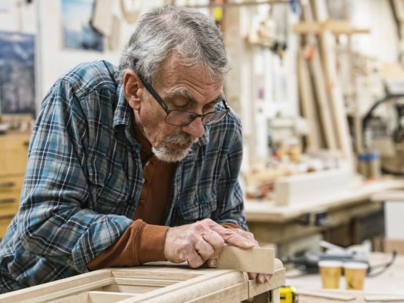 Senior Caucasian  carepenter sanding a wooden cabinet part in a large woodworking shop.